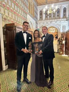 Da esquerda para direita: um homem alto de óculos de grau e terno preto, uma mulher de vestido preto longo e maquiagem de festa e um homem de terno preto e gravata borboleta. Todos segurando dois troféus.