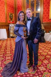 Da esquerda para direita: uma mulher de vestido preto longo e maquiagem de festa segurando o troféu e um homem de terno preto e gravata borboleta. Ambos sorrindo para a foto.