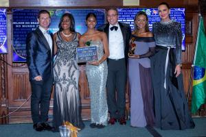 Da esquerda para direita: um homem vestindo terno azul marinho, uma mulher com um batom vermelho e cordão brilhoso e vestindo um vestido longo prateado, uma mulher vestindo um vestido longo prateado tomara-que-caia e segurando a placa de vencedora, um hom