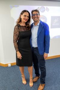 Da esquerda para direita: uma mulher de vestido preto e cabelo solto e um homem usando blazer azul e óculos de grau.