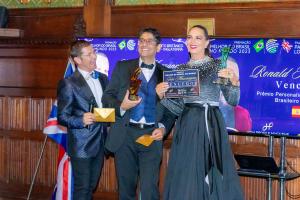 Da esquerda para direita: um homem vestindo terno azul marinho, outro homem segurando o troféu e usando gravata borboleta, terno e colete e uma mulher segurando a placa do vencedor e vestindo um vestido longo de festa.