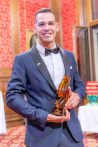 Um homem vestindo terno azul e gravata borboleta e segurando o troféu com as duas mãos. O fundo da imagem é um ambiente em tom vermelho.