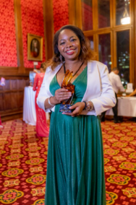 Uma mulher usando um vestido verde, sorrindo radiante enquanto segura um troféu. Seus cabelos castanhos estão presos para trás. A mulher parece orgulhosa e feliz por estar segurando seu prêmio. Olhando diretamente para a câmera com uma expressão de alegri