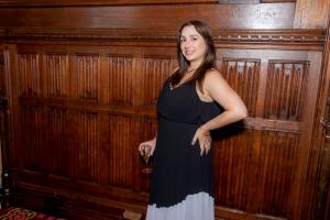 Uma mulher de cabelos castanhos fazendo uma pose lateral com a mão esquerda na cintura e a direita segurando uma taça. Ela usa vestido preto com a barra cinza e o modelo de alças. Ela sorrir para a foto.
