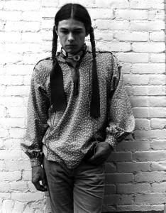 Black and white photograph of Native American subject, Rick Williams, leaning against a brick wall, part of the James Bama Collection in the McCracken Research Library at the Buffalo Bill Center of the West