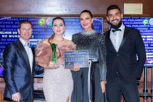 Da esquerda para direita: um homem de terno azul marinho escuro e usando óculos de grau e uma gravata borboleta, uma mulher de vestido de festa rose segurando com uma mão um troféu verde e com a outra uma placa de vencedora, uma mulher alta de vestido pre