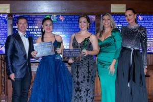 Da esquerda para direita: um homem de terno azul marinho e gravata borboleta usando óculos de grau, ao lado uma mulher de vestido de festa de alça azul marinho segurando uma placa de vencedora e um troféu, uma mulher vestindo um vestido de festa de alça p