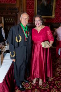 Da esquerda para direita: um homem de roupa preta e com algumas medalhas no pescoço, usa óculos de grau e tem cabelos brancos e uma mulher de cabelo bem curto estilo chanel usando um vestido vermelho longo de festa.