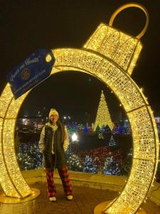 Woman wearing winter clothing posing under a giant lighted walk-through ornament Christmas decoration with Christmas trees and lights in the background.