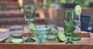 An image of handmade green glassware from Intertwined: Handmade for Good's new collection. From left to right, there is an old-fashioned green glass, a green wine glass, a green martini glass, a green stemless wine glass, a green champagne flute, and a gr