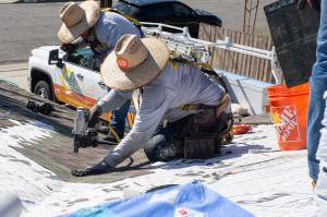 Two roofers install composite shingles on the roof of a one story house. There is an HES Solar work truck in the background