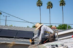 A roofer nails composite shingles onto a roof. There are three tall palm trees in the background.