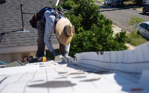 One roofer bends over and work on a single story home's roof. The roof in the foreground is covered with new roofing paper awaiting new shingles.