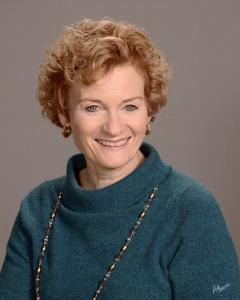 Sue Lusi poses for a headshot in front of a gray background. She is smiling and wearing a teal green turtleneck sweater.