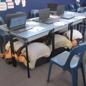 Photo showing 6 children beneath their desks bent over and covering their heads while holding onto legs of the desk.
