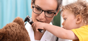 Child looking in a stuffed bear's ear with an otoscope while an audiologist supervises