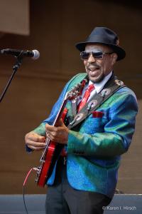 John Primer in an irradescent blue jacket playing his red guitar at the Chicago Blues Festival 2023
