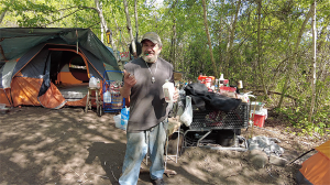 Homeless man in woods, where he lives in a tent, standing.