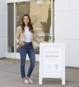 Photo of Courtney Claghorn, co-founder of SUGARED + BRONZED, standing by a sidewalk sign in front of a location of sugared + bronzed in CA