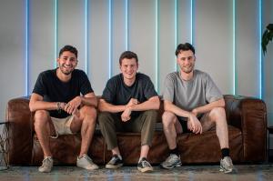 Three young men (the founders of Ivy The founder of Ivy [left-right - Gilad Bechor, CPO, Niklas Terrahe, CEO, Liam Hänel, COO]) sitting on a brown couch together looking really happy. The background is grey with colourful stripe lighting and a plant off t