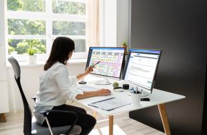 A health practitioner utilizing the Power Diary practice management software on dual monitors in a sunlit, contemporary workspace. While one screen displays a colorful calendar schedule, the other presents detailed client data. The professional setting em
