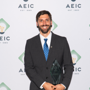 Andrew is in a black suit with a white dress shirt and blue tie. He is holding an AEIC trophy in his hand and smiling at the camera. He is standing in front of a step and repeat backdrop with the AEIC logo on it.