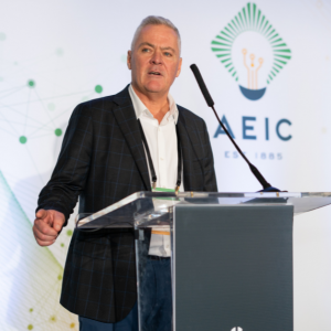 Steve is in a black blazer and white dress shirt. He is talking at the podium and the green and yellow AEIC logo is on the screen behind him.