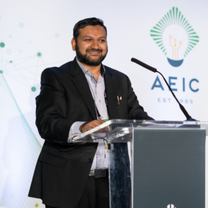 Umair is smiling at a podium. He is wearing a black blazer with a pin-striped dress shirt. He has black hair and a beard. There is a green and yellow AEIC logo behind him on a screen.