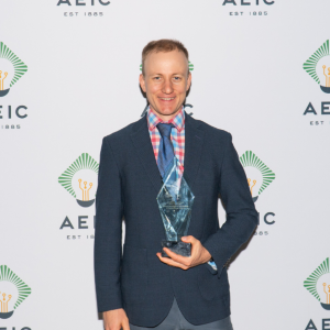 Vlad is holding an AEIC trophy in his hand. He is wearing a navy blazer with a red and blue checkered dress shirt and blue tie. He is standing in front of a step and repeat backdrop that has the AEIC logo on it.