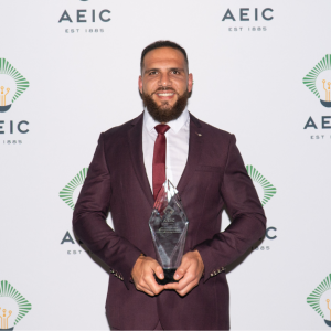 Mohammad is holding an AEIC trophy in his hands. He is wearing a maroon suit with a white dress shirt and red tie. He is standing in front of a step and repeat backdrop that has the AEIC logo on it.