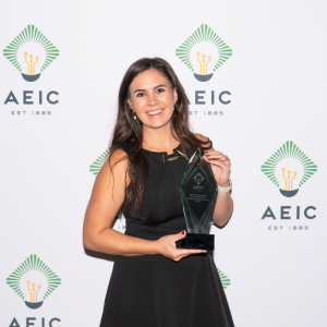 Sarah is holding an AEIC trophy in her hands. She is wearing a black, sleeveless dress. She has long, brown hair and brown eyes. She is standing in front of a step and repeat backdrop that has the AEIC logo on it.