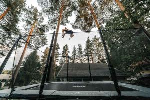 A man is doing a high backflip on a large black, rectangular trampoline, surrounded by pine trees.