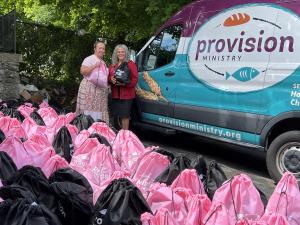 Two women smiling for camera holding black and pink drawstring bags. Van in image with "Provision Ministry" written with cartoon bread and fish surrounding the words.
