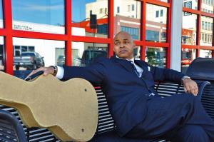 Image of Author/guitarist Maurice Johnson sitting on an Oklahoma City downtown bench in a blue suit with a guitar.
