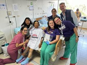 A grandmother who just received a knee replacements sits on the edge of her hospital bed surrounded by her granddaughter and the Operation Walk Maryland volunteer medical team. Niza is sporting a shirt with the organization's logo.
