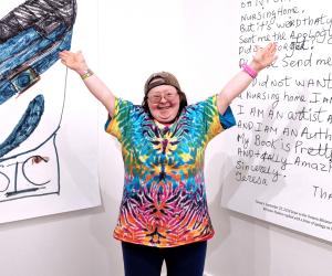 Teresa Heartchild, a woman with Down Syndrome, wearing a tie-dye t-shirt. She stands in front of her large art posters in an art gallery in Vancouver.