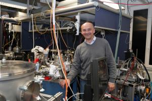 A man stands in a laser lab surrounded by cables, tubes, machines and optical tables.