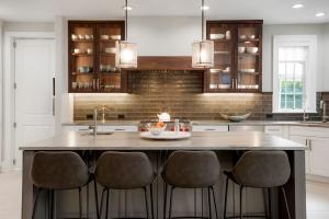 A bright and modern kitchen featuring a large island with four chairs, a golden brick backsplash, brown open faced cabinets.