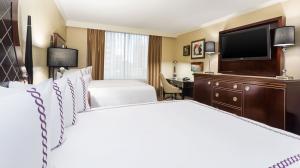 Elegant hotel room with two beds, crisp linens, classic furniture, and a large TV, bathed in natural light.