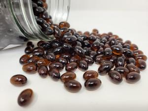 A glass jar with thousands of golden and dark gelcaps spilling out, jar on its side, gelcaps rolling out of it.