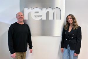 Rob Matlow and Jessica Chalk standing with the REM logo between them.