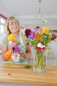 An executive housekeeper with glasses and a floral apron arranging a colorful bouquet of fresh flowers in a clear vase on a wooden kitchen table, preparing a decorative floral arrangement for her employer's home