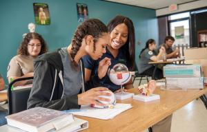 A program instructor and participant are seated in a classroom looking at plastic models of an eye and an ear. There are three students in the background seated at their desks.