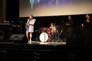 A band performs on stage at The Strand Theatre, featuring a young female saxophonist in the foreground, a boy on drums, and two men, one playing the clarinet and the other the guitar, under a blue-lit backdrop.