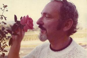 A vintage photograph of Norman Blagman, with a mustache and curly hair, smiling as he gently smells a pink rose held in his hand, with a blurred background suggesting a vineyard.