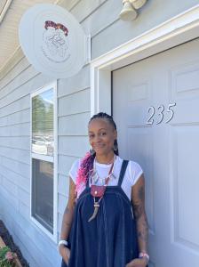 smiling midwife standing by a blue door with numbers 235 and Gather Grounded Midwifery sign overhead
