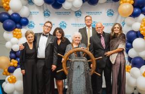 Selz Family at  Thomas House Family Shelter Autumn Reflections Gala posing with ship's wheel at gala event, surrounded by blue, white, and gold balloons, representing their guidance and steadfast direction in support of unhoused families in need.