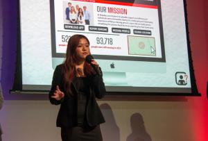 This image shows a high school student onstage with a microphone. A slide from her team's pitch presentation is on the screen behind her.