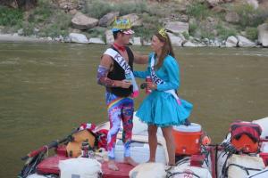 A bachelor and bachelorette wearing king and queen sashes stand on a whitewater rafting boat and smile at each other during their joint bachelor and bachelorette river trip