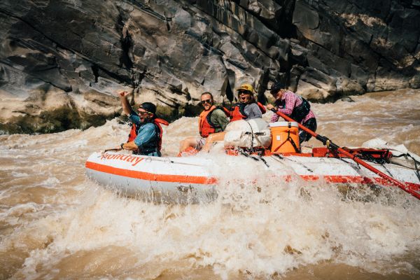 Guide rows three guests through a large rapid in Westwater Canyon. The guests are sitting in the front of the raft and are splashed with water while smiling and cheering.